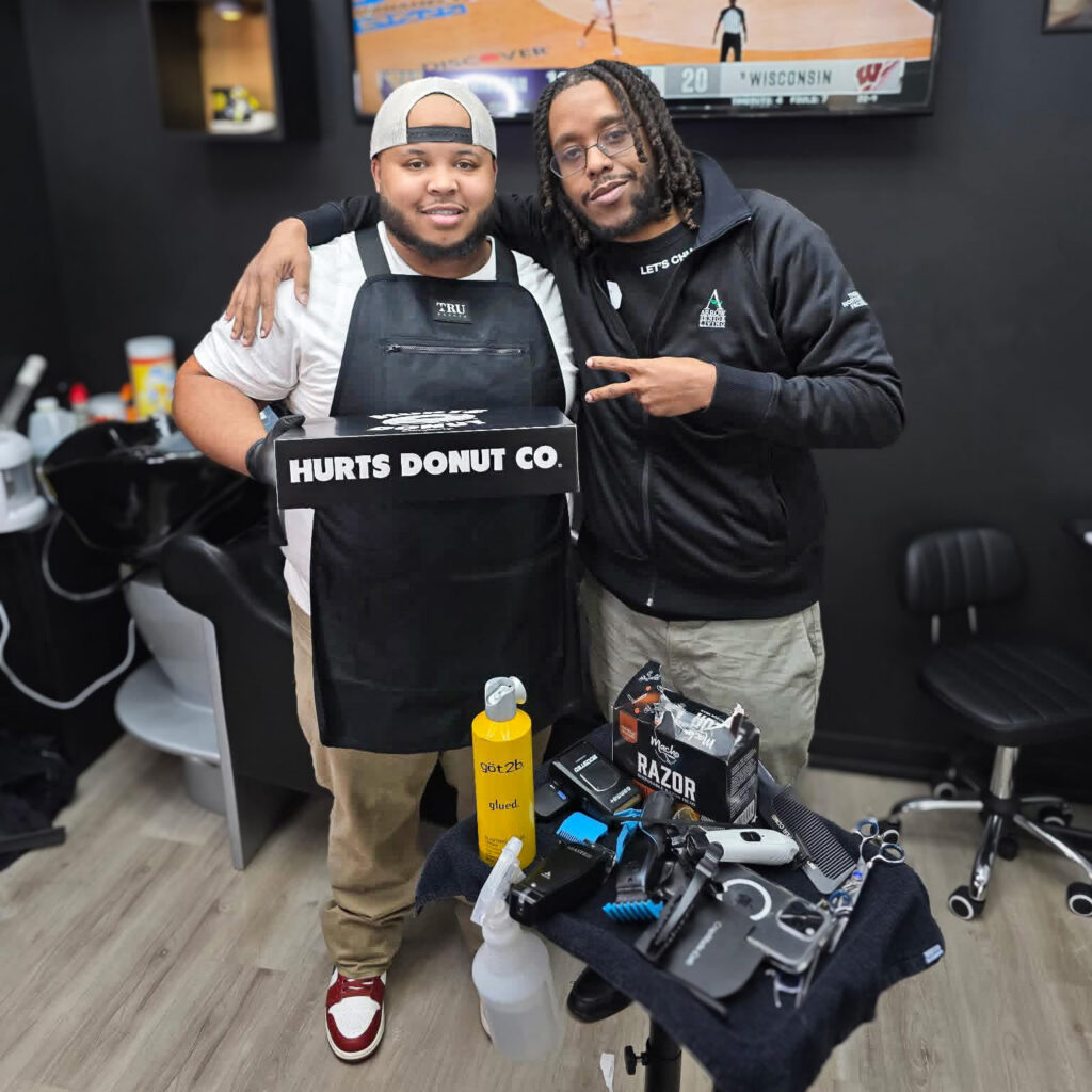 The owner of a local barbershop, FadezbyBoda, smiles alongside a team member from Walden Place Senior Living, holding a gifted box of doughnuts.