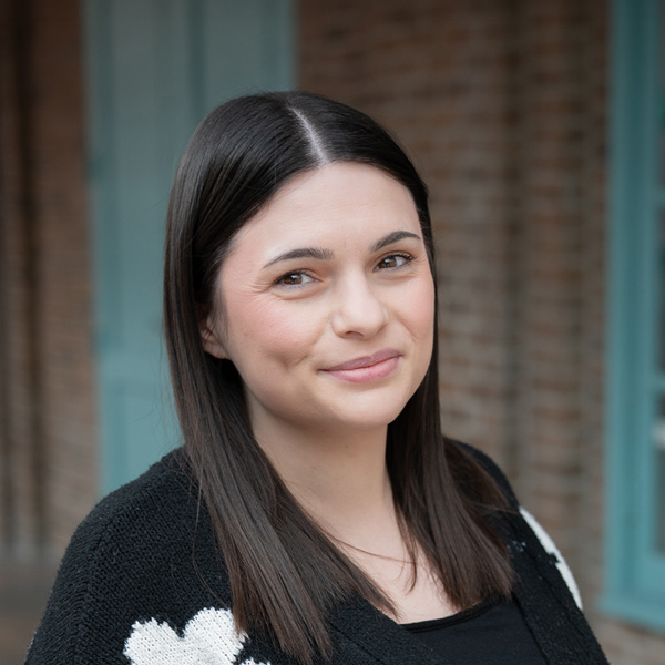 Emily Thompson, Resident Services Director at Walden Place Senior Living, smiling in a professional headshot, wearing a black top and patterned cardigan, with a softly blurred outdoor background featuring brick and teal architectural details.