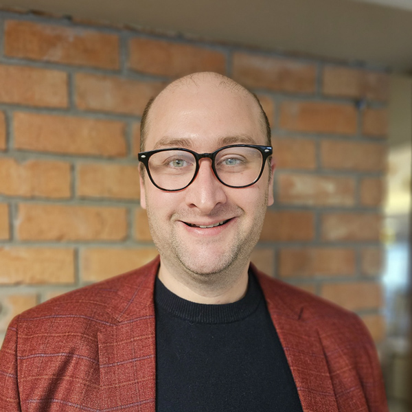 Josh Figg, Senior Living Director at Walden Place Senior Living, smiling in a professional headshot, wearing glasses, a black shirt, and a red blazer, with a softly blurred brick background.