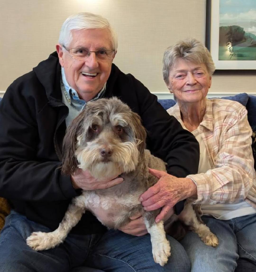 Two senior residents smile while sitting with their gray and white dog in a pet-friendly senior living community.