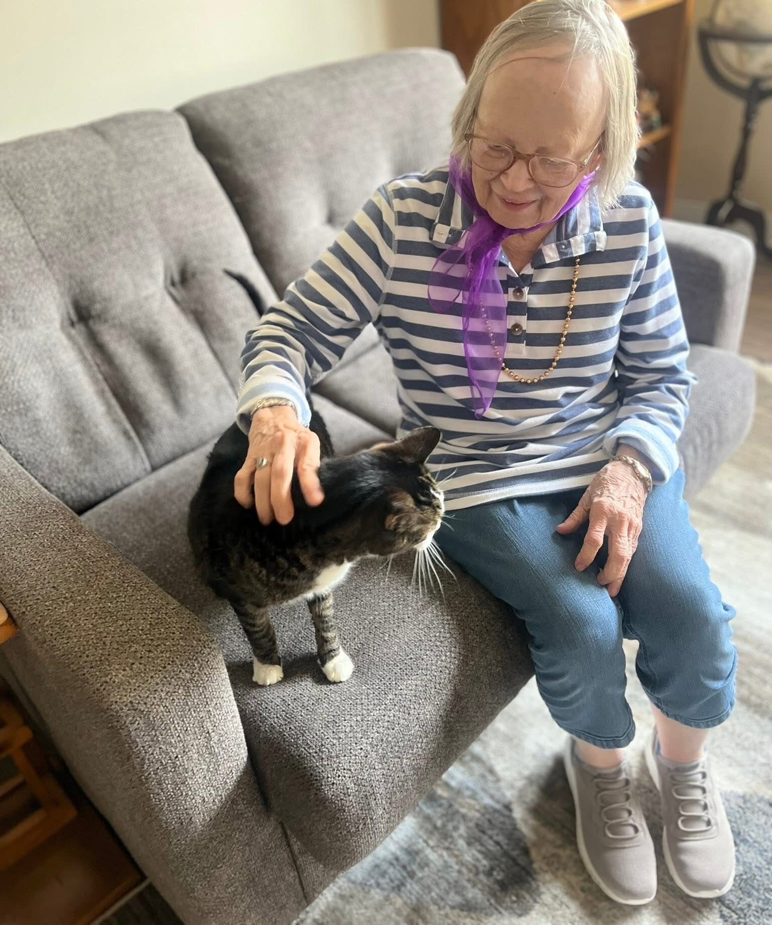 Senior resident smiles while petting a black and white cat on a gray couch in her senior living apartment.