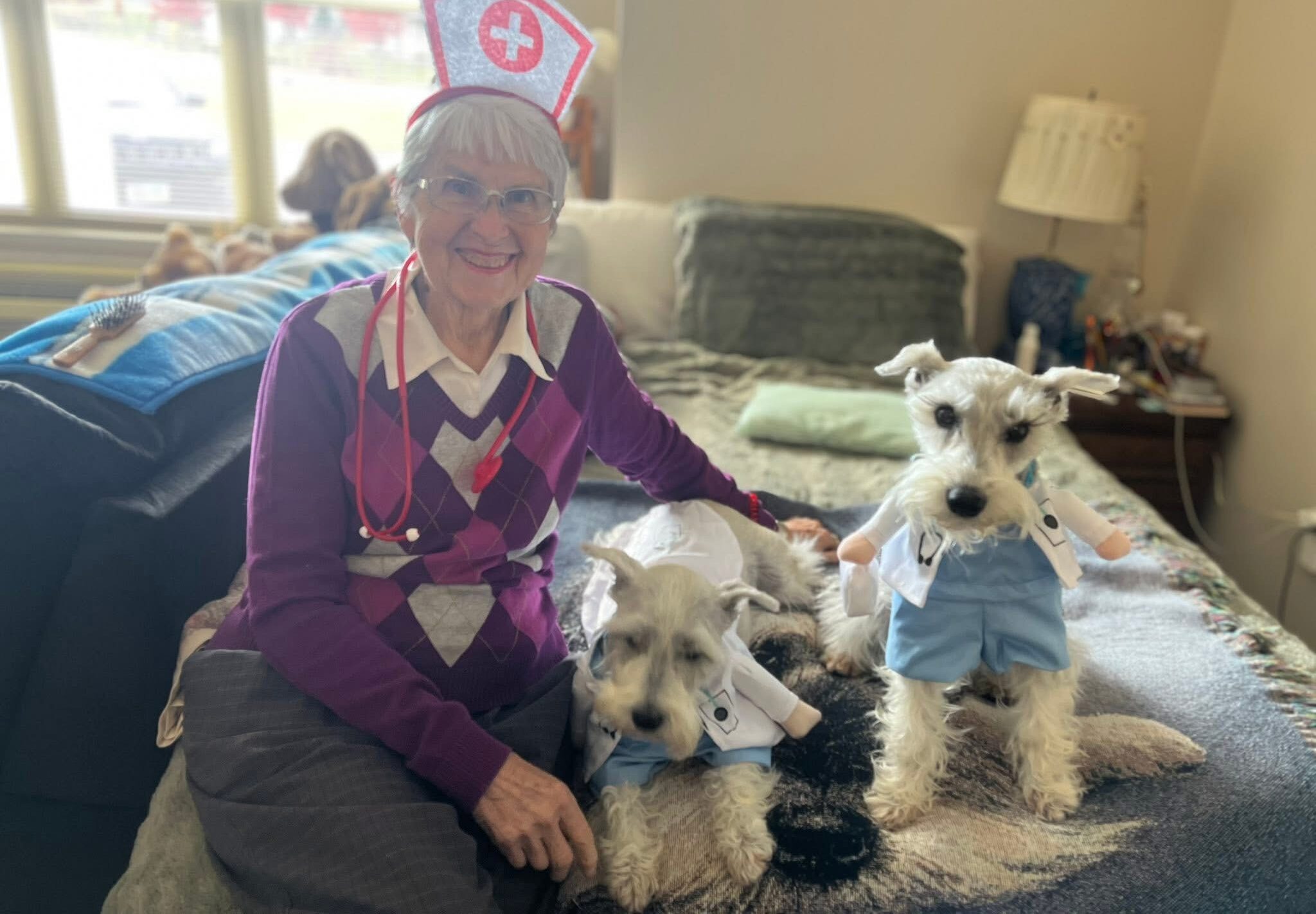 Senior resident wearing a playful nurse costume sits with two dressed-up dogs on a bed in a pet-friendly senior living apartment.