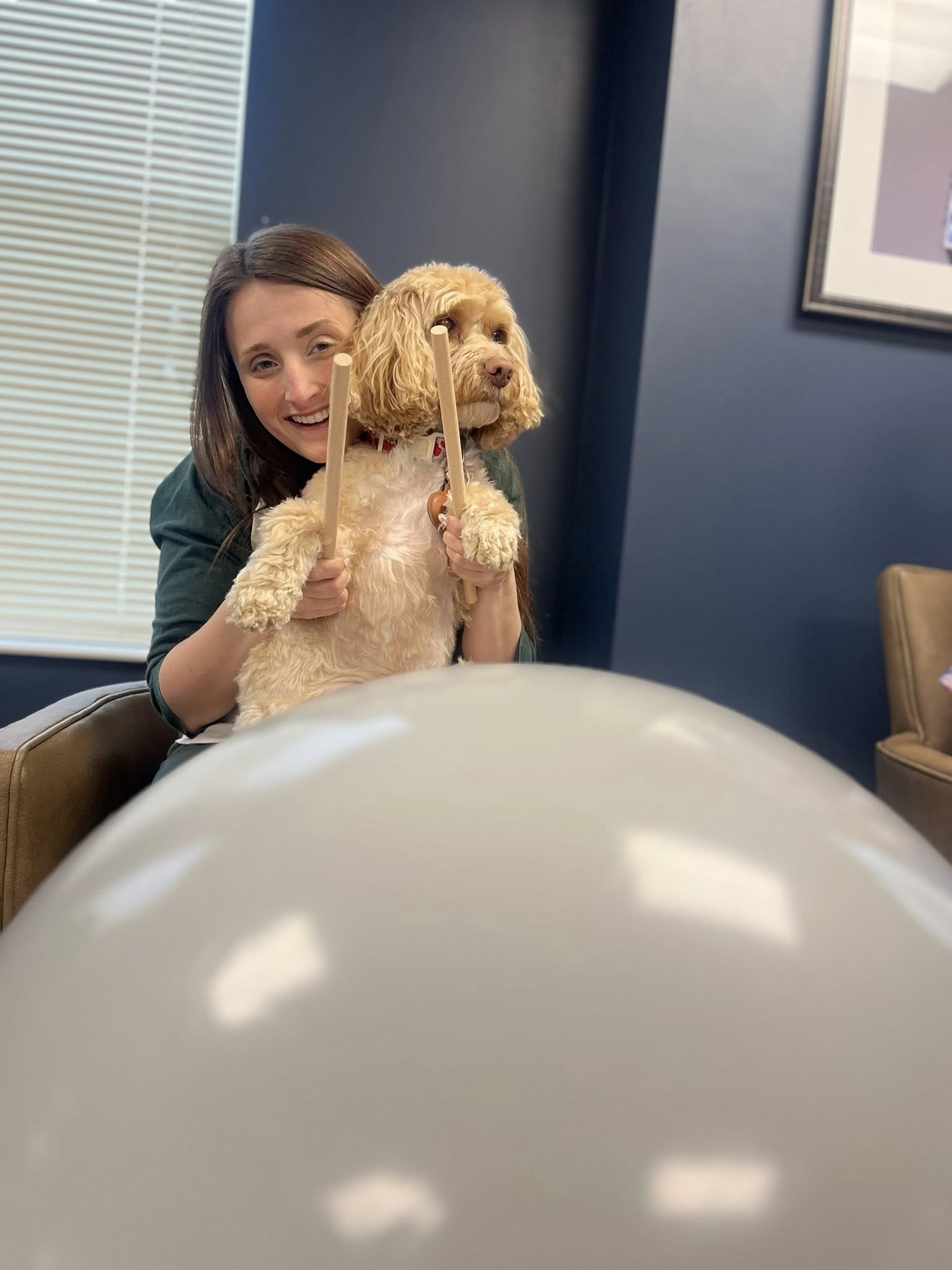 Team member helps a dog hold drumsticks during a playful pet-friendly moment in a senior living community.