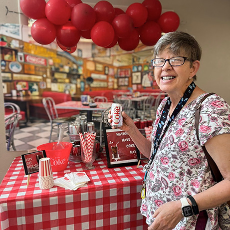 A resident at Walden Place smiles, holding a can of soda, in front of an event display with balloons, backdrop, and table filled with treats.